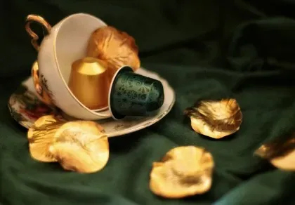 A tilted teacup with golden and green coffee capsules spills onto a dark green fabric, surrounded by golden leaves